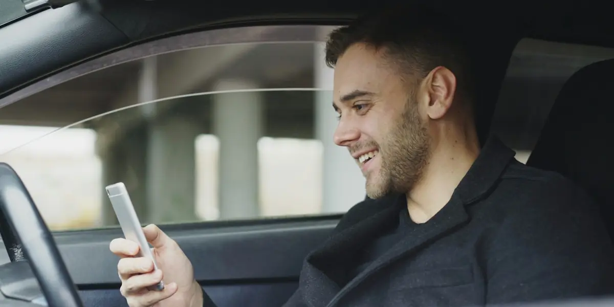 Smiling driver in his parked car, sending a request to a mobile mechanic from his phone